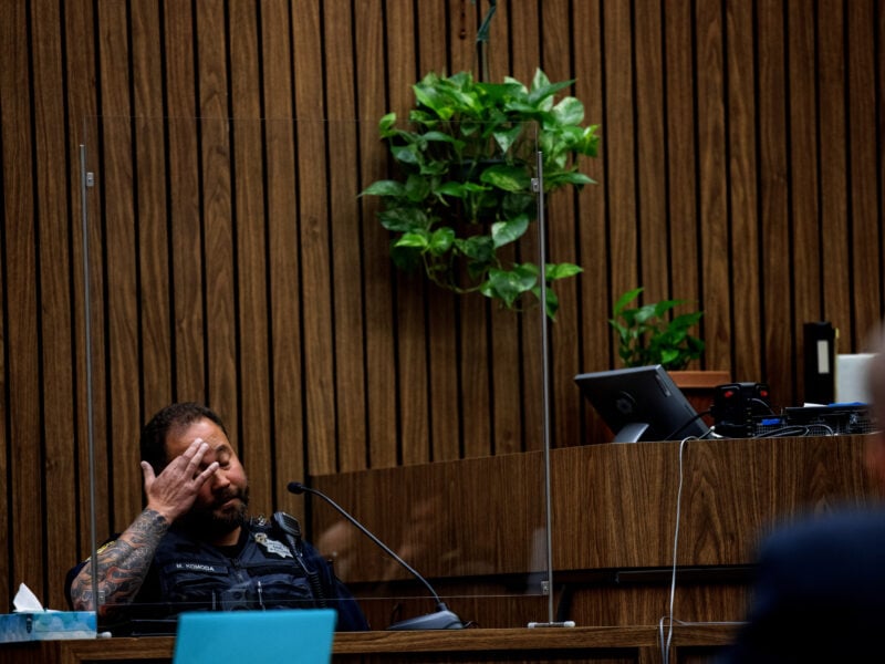 A uniformed police officer with visible arm tattoos sits behind a transparent barrier on a courtroom witness stand. The wood-paneled wall behind him features hanging green plants, and courtroom equipment is visible nearby.