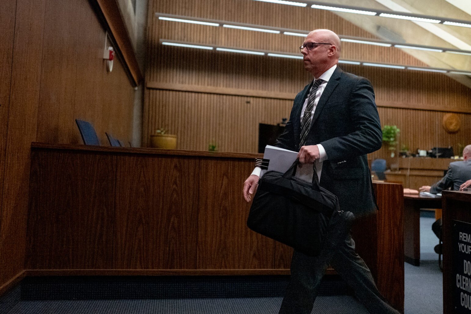 A man in a dark suit and tie, carrying a dark bag and court documents, walks briskly past the witness stand inside a dimly lit courtroom with wood-paneled walls and vaulted ceiling beams.
