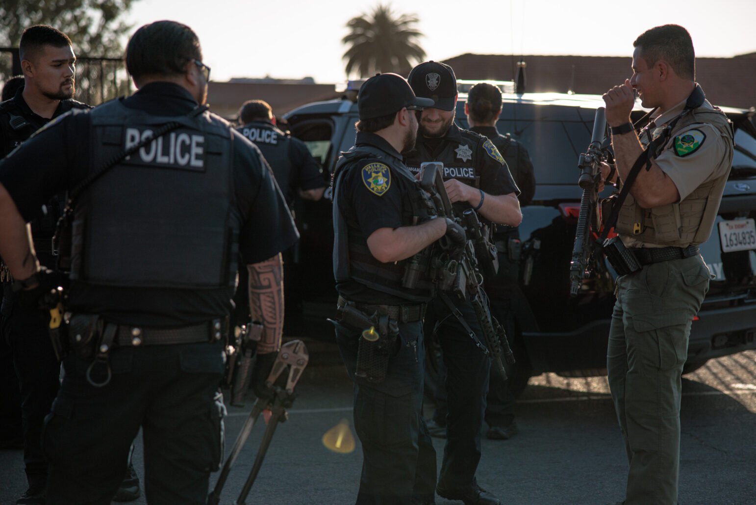 Multiple armed officers in tactical vests stand near a black police SUV in daylight, preparing for a coordinated operation. Several of the officers are carrying rifles. The setting appears to be a perimeter area, with the sun casting long shadows.