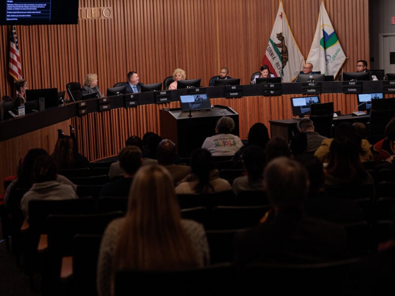 Vallejo City Council meeting in session, with council members seated at a long curved dais, facing an audience of community members in the council chamber. California and city flags are displayed behind the council.