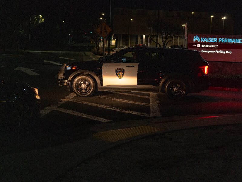 A Vallejo police SUV leaves the Kaiser Permanente emergency entrance at night. The illuminated hospital sign reads “EMERGENCY – Emergency Parking Only,” and directional road markings are visible under the vehicle.