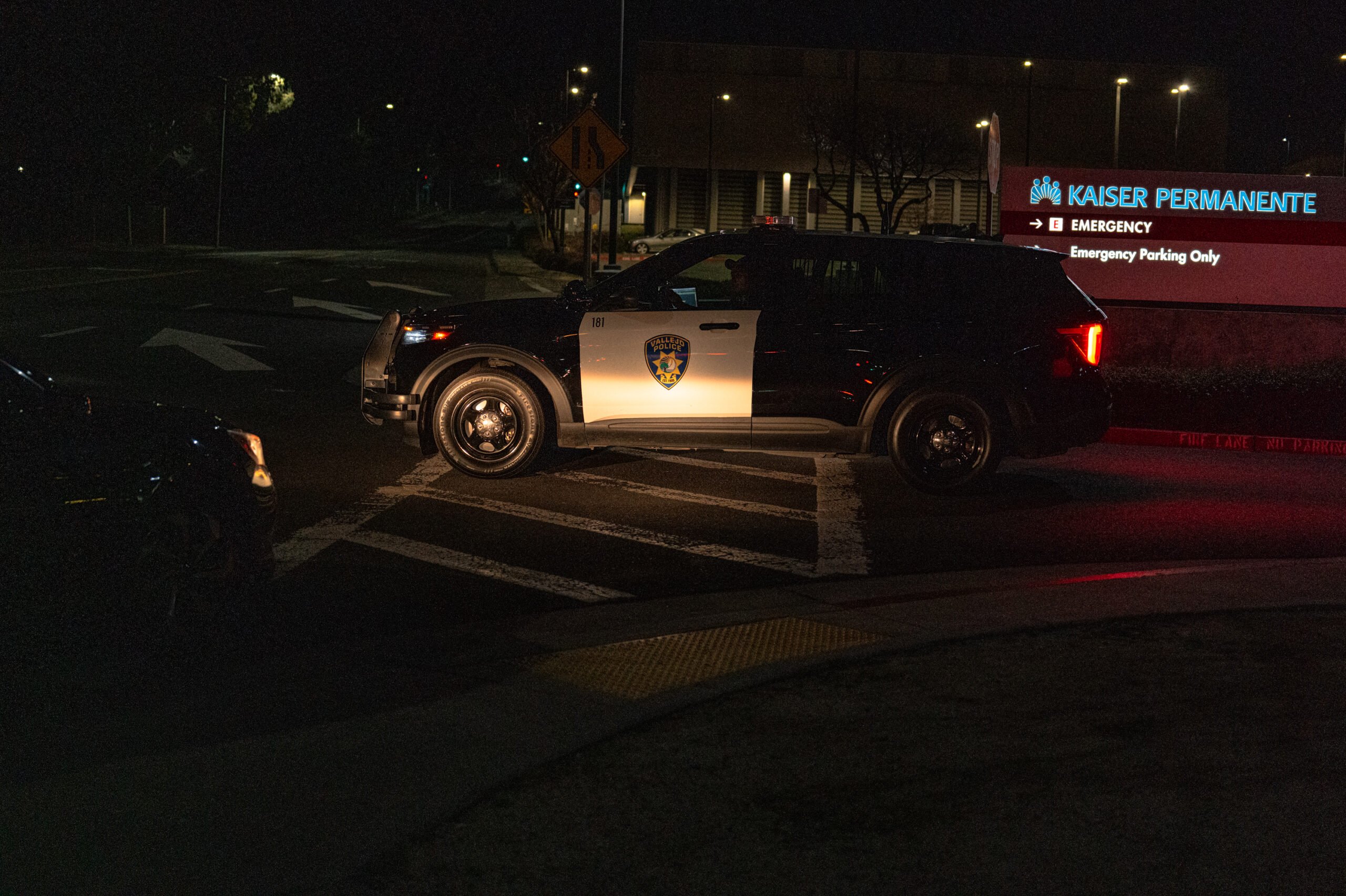 A Vallejo police SUV leaves the Kaiser Permanente emergency entrance at night. The illuminated hospital sign reads “EMERGENCY – Emergency Parking Only,” and directional road markings are visible under the vehicle.