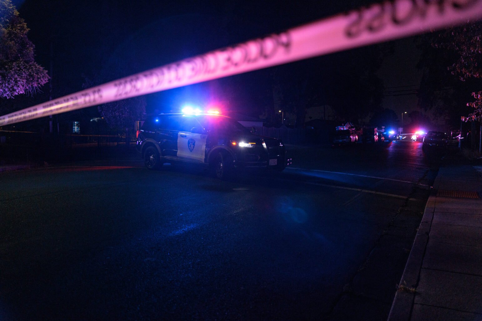 A Vallejo police SUV with flashing lights blocks a dark residential street behind yellow crime scene tape. Several other emergency vehicles with flashing lights are visible in the distance, casting blue and red reflections on the pavement.