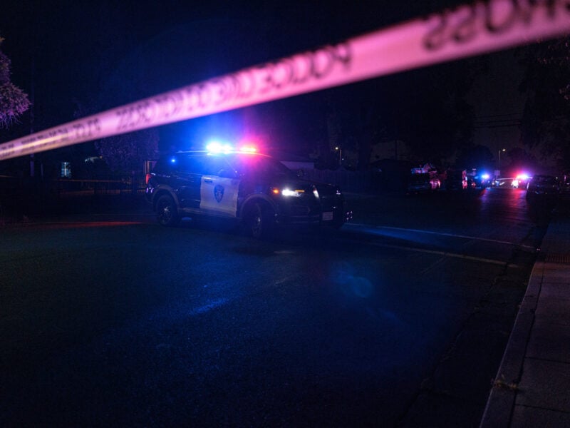 A Vallejo police SUV with flashing lights blocks a dark residential street behind yellow crime scene tape. Several other emergency vehicles with flashing lights are visible in the distance, casting blue and red reflections on the pavement.