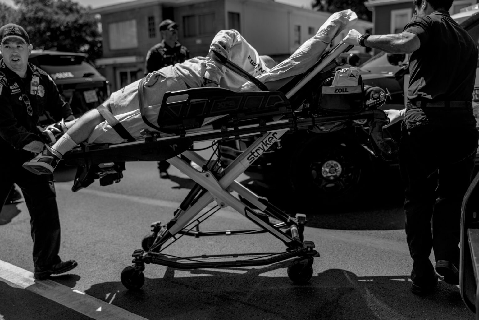 Emergency responders wheel a person strapped to a gurney across the street. The person is wearing light-colored pants and is partially covered with a sheet. Multiple police officers are visible in the background.