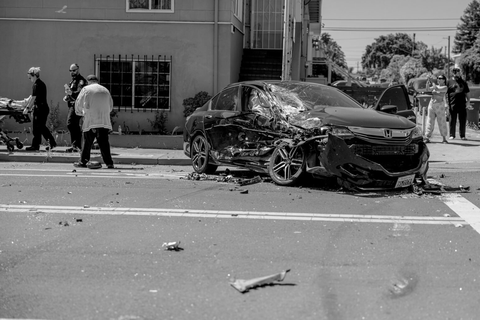 A damaged black Honda sedan rests in the street following a crash, its front end caved in and windshield shattered. Police officers, medics, and bystanders stand nearby, while debris litters the road.