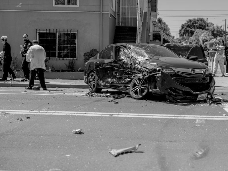 A damaged black Honda sedan rests in the street following a crash, its front end caved in and windshield shattered. Police officers, medics, and bystanders stand nearby, while debris litters the road.
