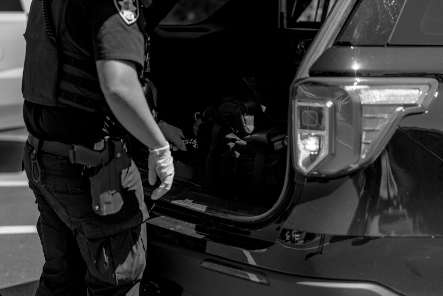 A police officer wearing gloves opens the rear of a patrol SUV, revealing gear and a bag inside. The officer's gun and radio are visible on their belt.