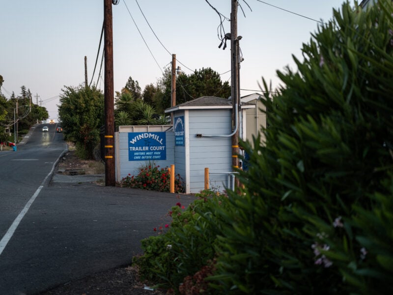 A street view shows the entrance to Windmill Trailer Court at dusk. A blue sign reads, "Windmill Trailer Court. Visitors must park cars outside court." Power poles and lush greenery frame the entrance, while vehicles travel on the road in the distance.