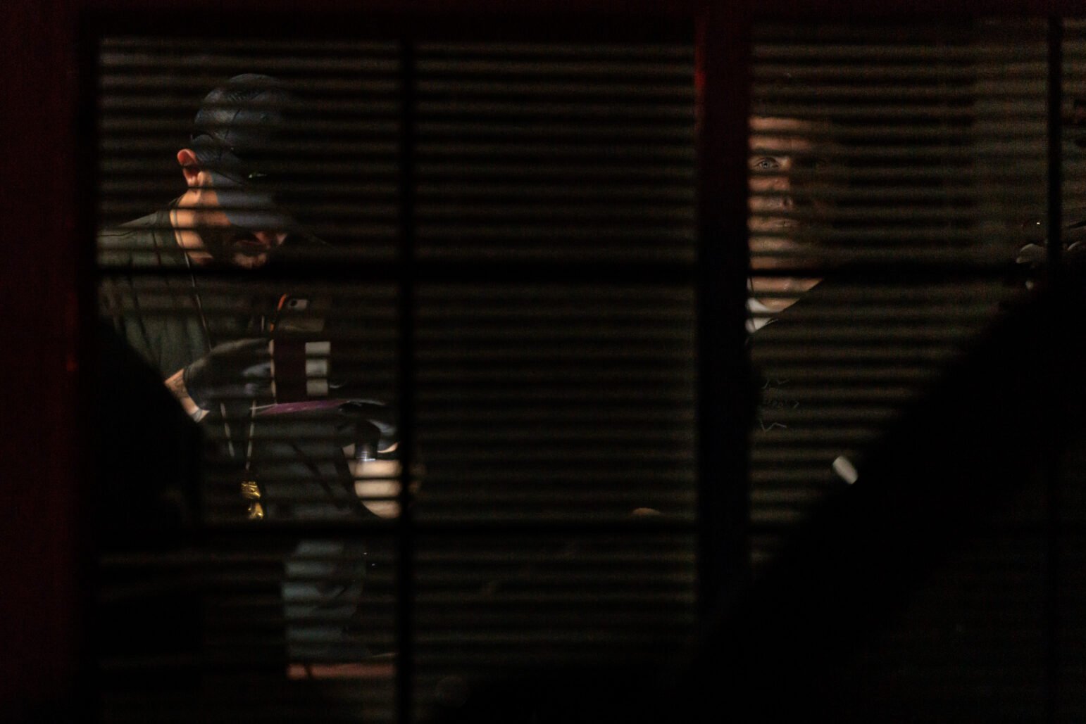 Two men, one wearing gloves and a lanyard with a badge, are dimly visible through partially closed window blinds at night. One man examines an object under a light, while the other looks out the window, his face partially illuminated in the darkness.