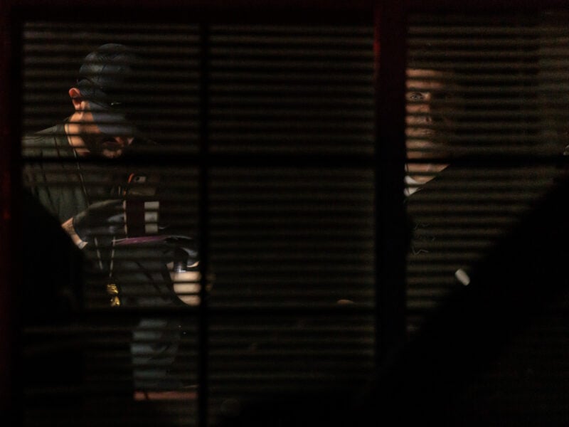 Two men, one wearing gloves and a lanyard with a badge, are dimly visible through partially closed window blinds at night. One man examines an object under a light, while the other looks out the window, his face partially illuminated in the darkness.