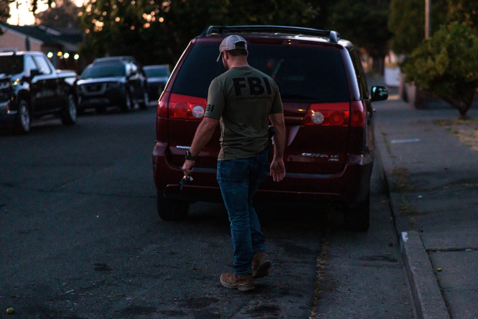 An FBI agent wearing a green t-shirt and baseball cap walks past a red SUV parked on a residential street at dusk, holding a set of keys in one hand. Several other vehicles are parked along the curb.