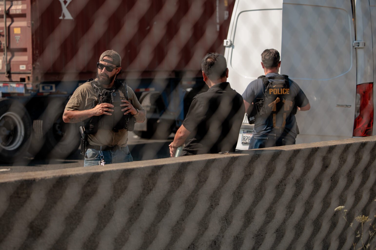Plainclothes officers stand near a white van along a freeway barrier. The scene is viewed through a chain-link fence.