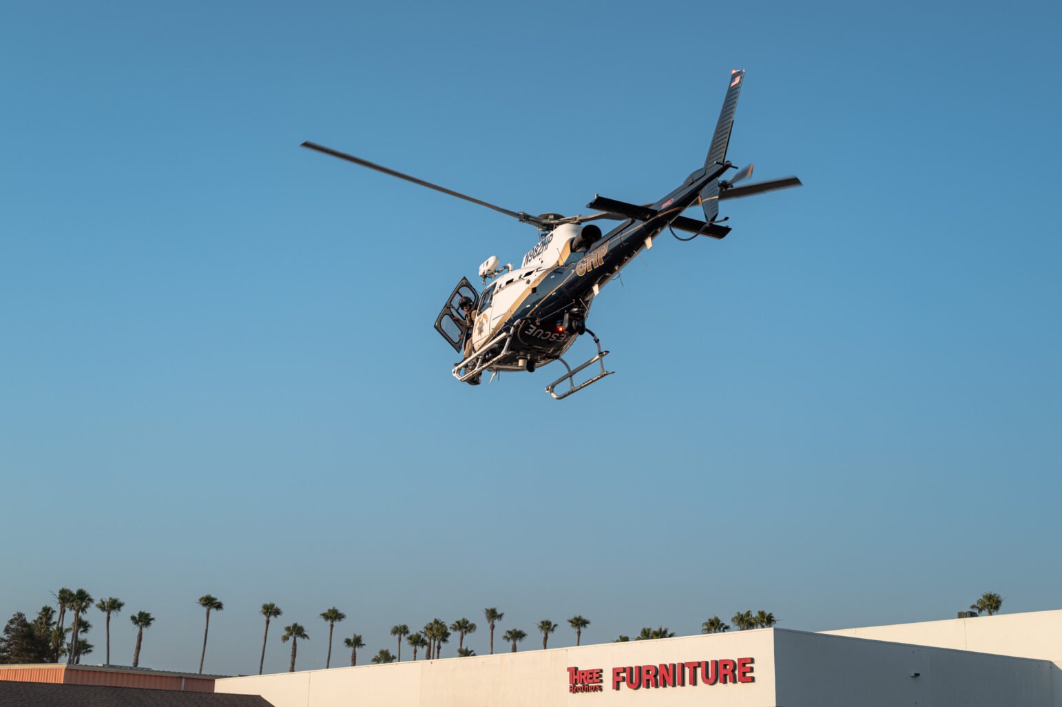 A California Highway Patrol rescue helicopter takes off over a strip mall with palm trees in the background. The aircraft’s door is open and a crew member is visible inside.