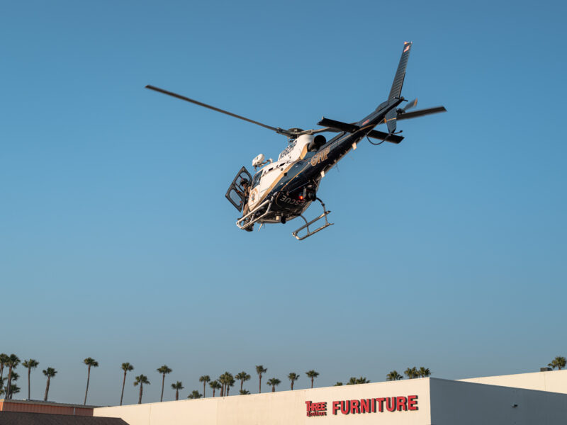A California Highway Patrol rescue helicopter takes off over a strip mall with palm trees in the background. The aircraft’s door is open and a crew member is visible inside.