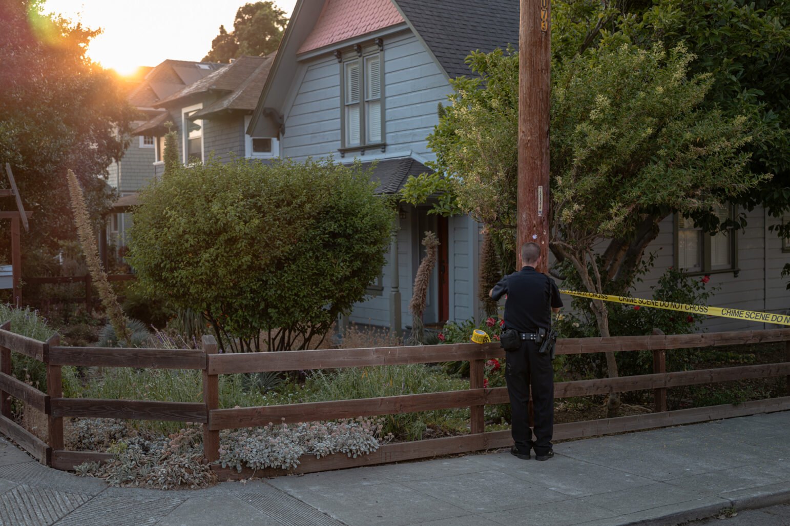 A Vallejo officer secures yellow crime scene tape to a utility pole outside a gray house with a wooden fence and well-kept garden, as the sun sets in the background.
