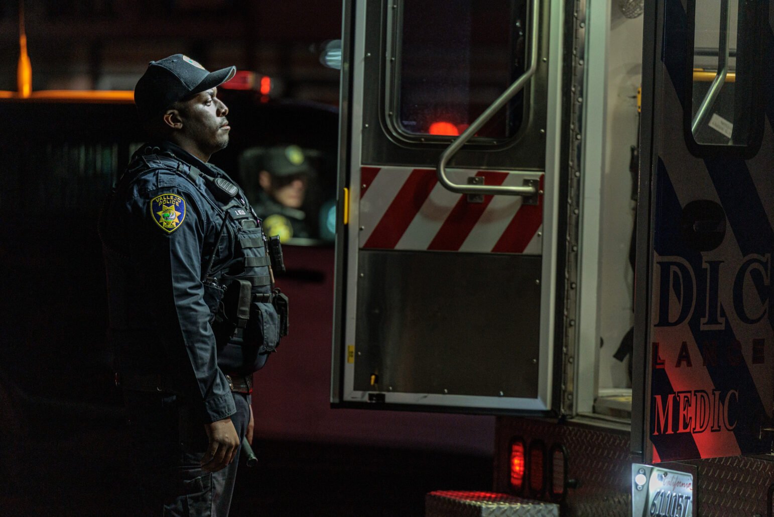 A Vallejo police officer in tactical gear stands solemnly beside an open ambulance door at night, illuminated by emergency vehicle lights. Another officer is visible seated in a nearby patrol car.