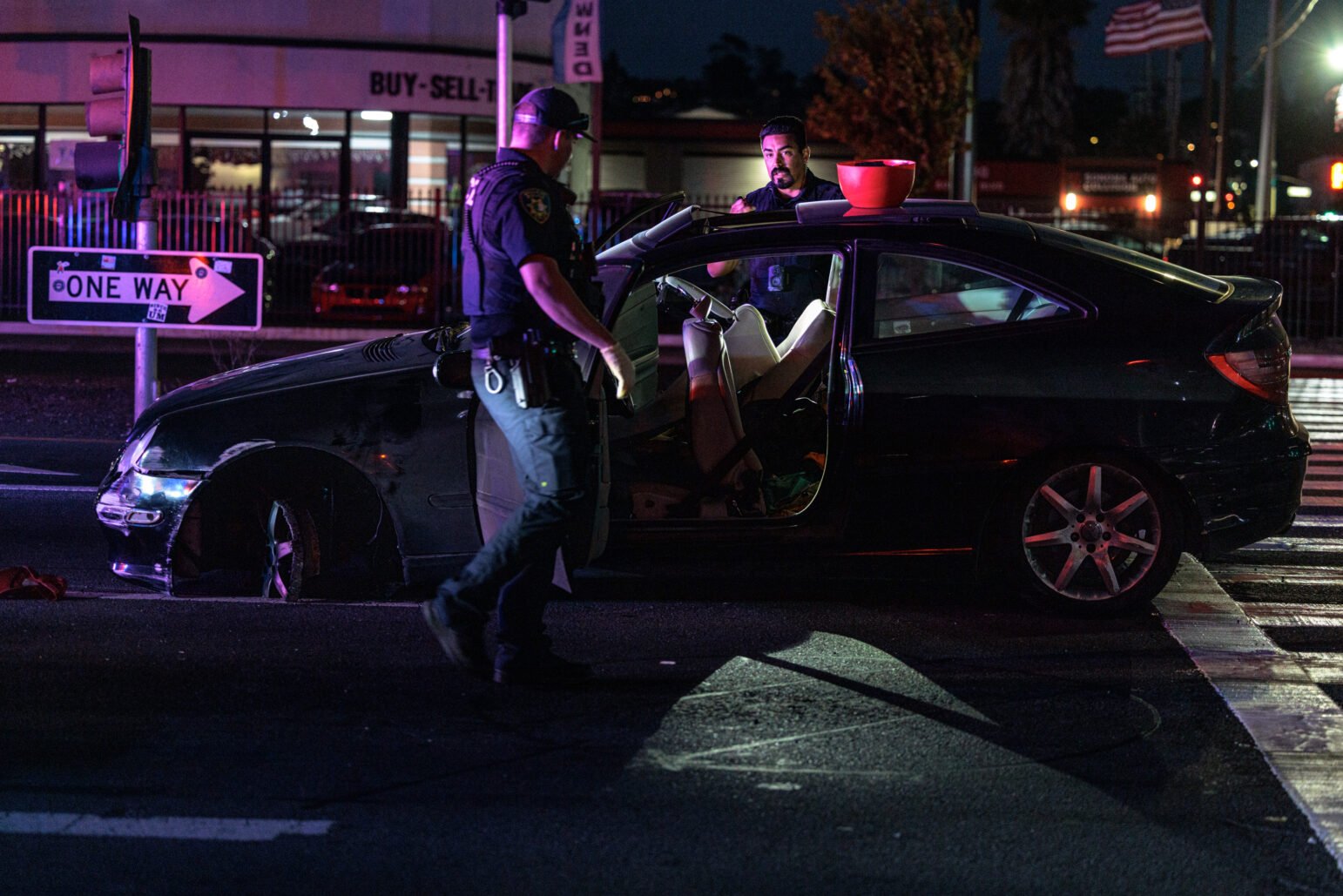 Two police officers investigate a heavily damaged vehicle stopped in an intersection at night. One officer stands beside the open car door while the other speaks to someone inside. A red plastic bowl rests atop the car roof, and surrounding businesses are lit in the background. A “One Way” traffic sign and an American flag are visible nearby.