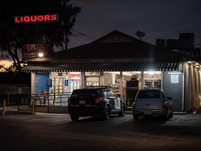 A Vallejo police SUV and a silver PT Cruiser are parked outside Val’s Wagon Wheel Liquors at dusk. The store’s bright interior contrasts with the darkened sky, and a red neon “LIQUORS” sign glows above the building.