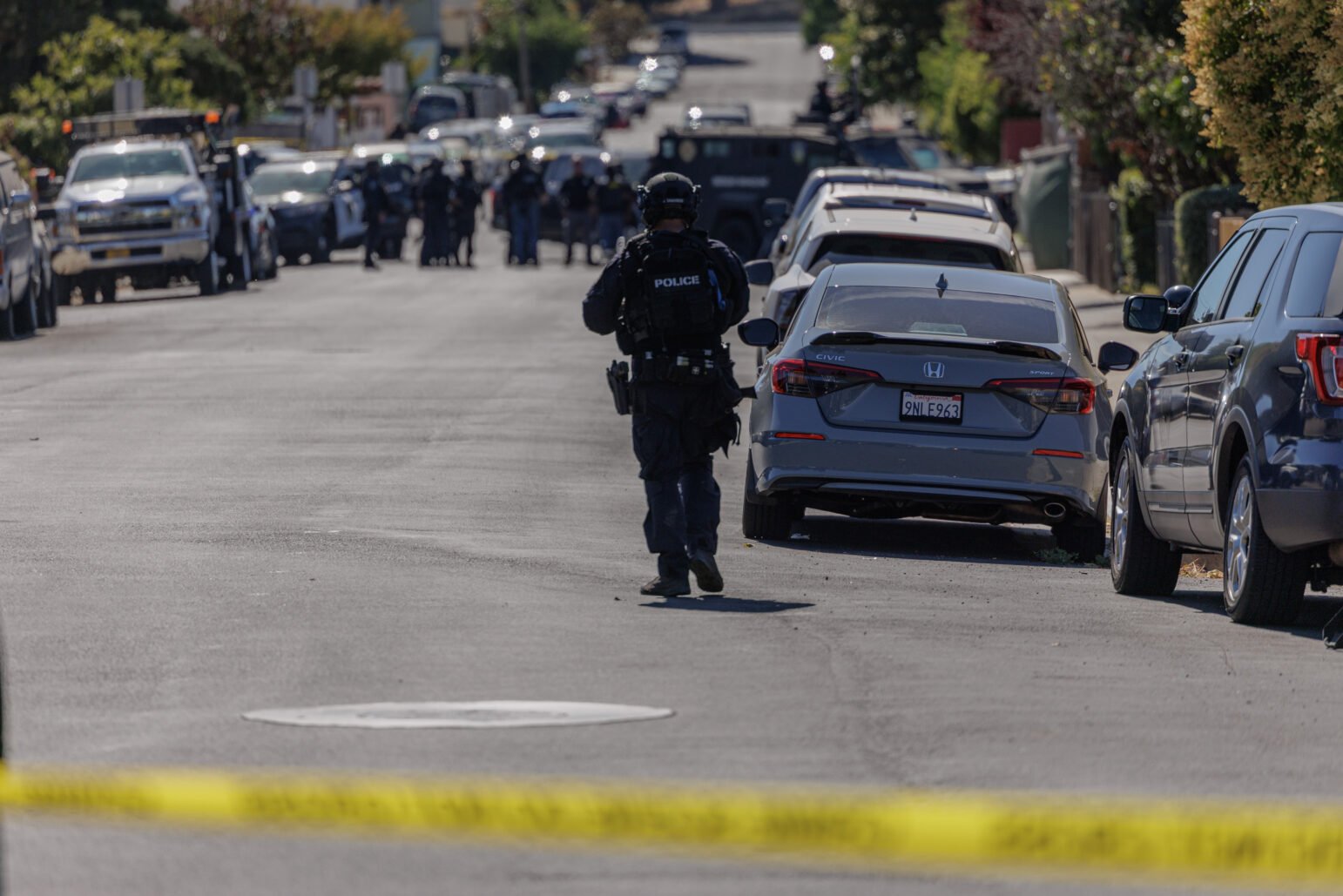 A police officer in tactical gear walks down a residential street cordoned off with yellow tape, approaching a group of officers and law enforcement vehicles in the distance, including an armored vehicle. Parked cars line both sides of the street under bright daylight.