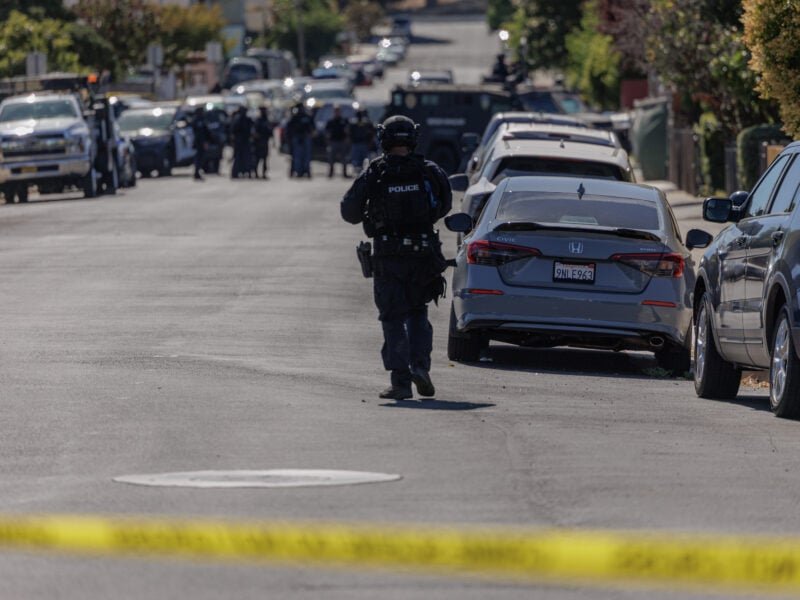 A police officer in tactical gear walks down a residential street cordoned off with yellow tape, approaching a group of officers and law enforcement vehicles in the distance, including an armored vehicle. Parked cars line both sides of the street under bright daylight.