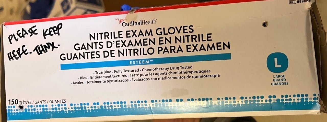A box of Cardinal Health nitrile exam gloves labeled in English, French, and Spanish is marked with a handwritten message in black ink: “PLEASE KEEP HERE. THNX.” The box contains 150 large-sized gloves and is slightly dented near the upper corner. Two small holes are visible in the side of the box.