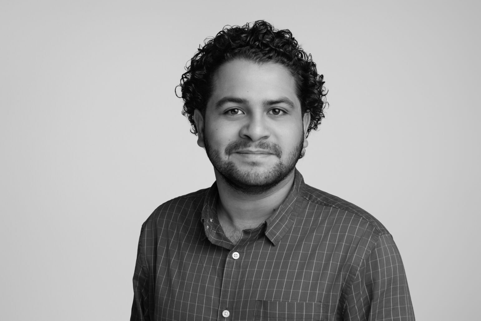 A black-and-white studio portrait of a man with short curly hair and a trimmed beard, wearing a checkered button-up shirt. He faces the camera with a neutral, slight smile against a plain light background.