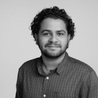 A black-and-white studio portrait of a man with short curly hair and a trimmed beard, wearing a checkered button-up shirt. He faces the camera with a neutral, slight smile against a plain light background.