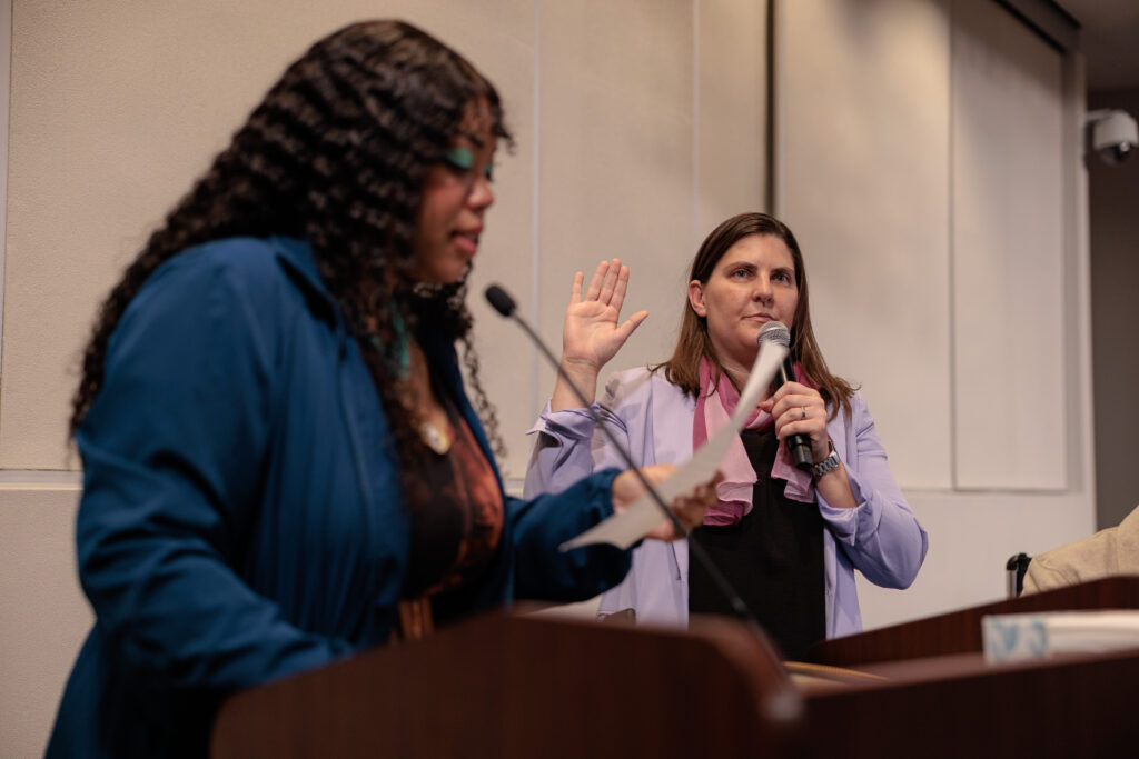 A woman in a light purple blazer stands with her right hand raised and holds a microphone in her left as she takes an oath, while another woman in the foreground reads from a paper at a podium. The setting appears to be an official indoor ceremony or city government event.