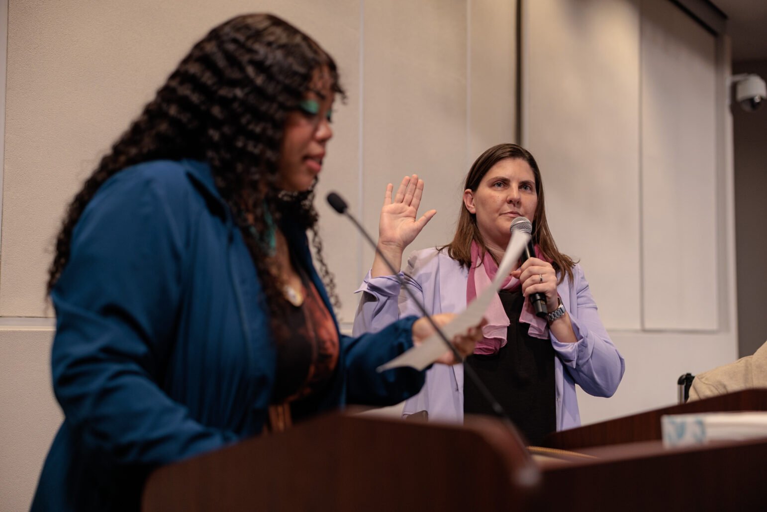 A woman in a light purple blazer stands with her right hand raised and holds a microphone in her left as she takes an oath, while another woman in the foreground reads from a paper at a podium. The setting appears to be an official indoor ceremony or city government event.