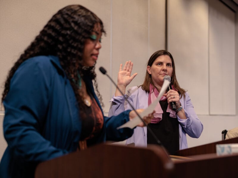A woman in a light purple blazer stands with her right hand raised and holds a microphone in her left as she takes an oath, while another woman in the foreground reads from a paper at a podium. The setting appears to be an official indoor ceremony or city government event.