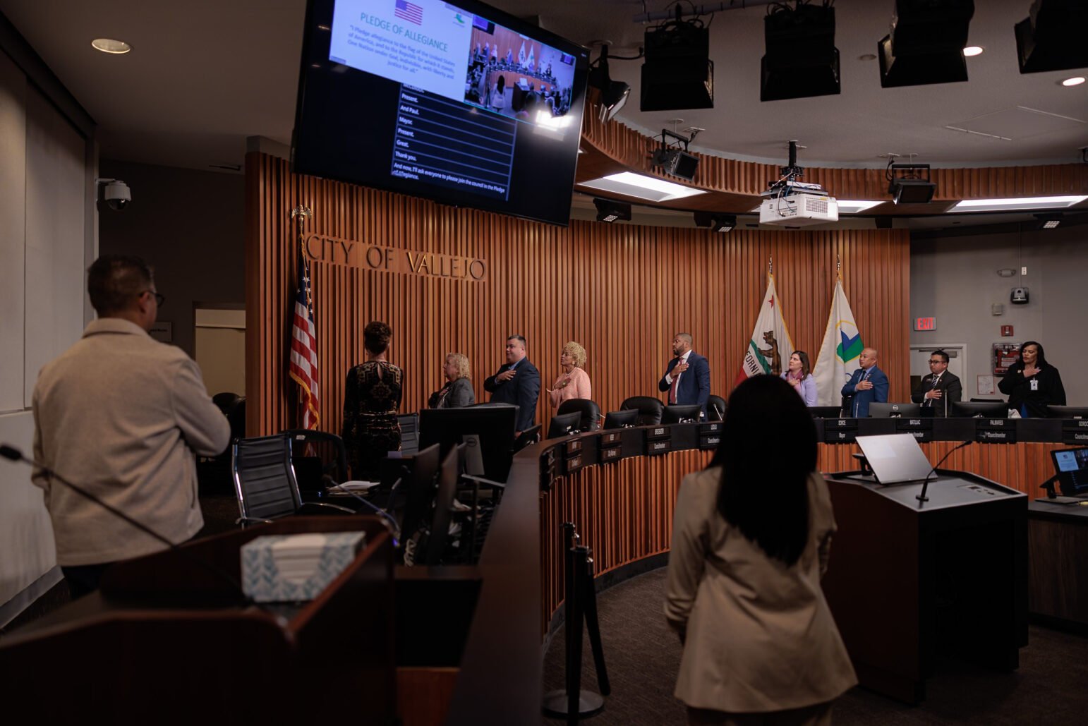 Members of the Vallejo City Council and attendees stand with hands over hearts during the Pledge of Allegiance inside the council chambers. The American flag, California state flag, and City of Vallejo flag are displayed behind the council dais. A large screen overhead displays the pledge text.