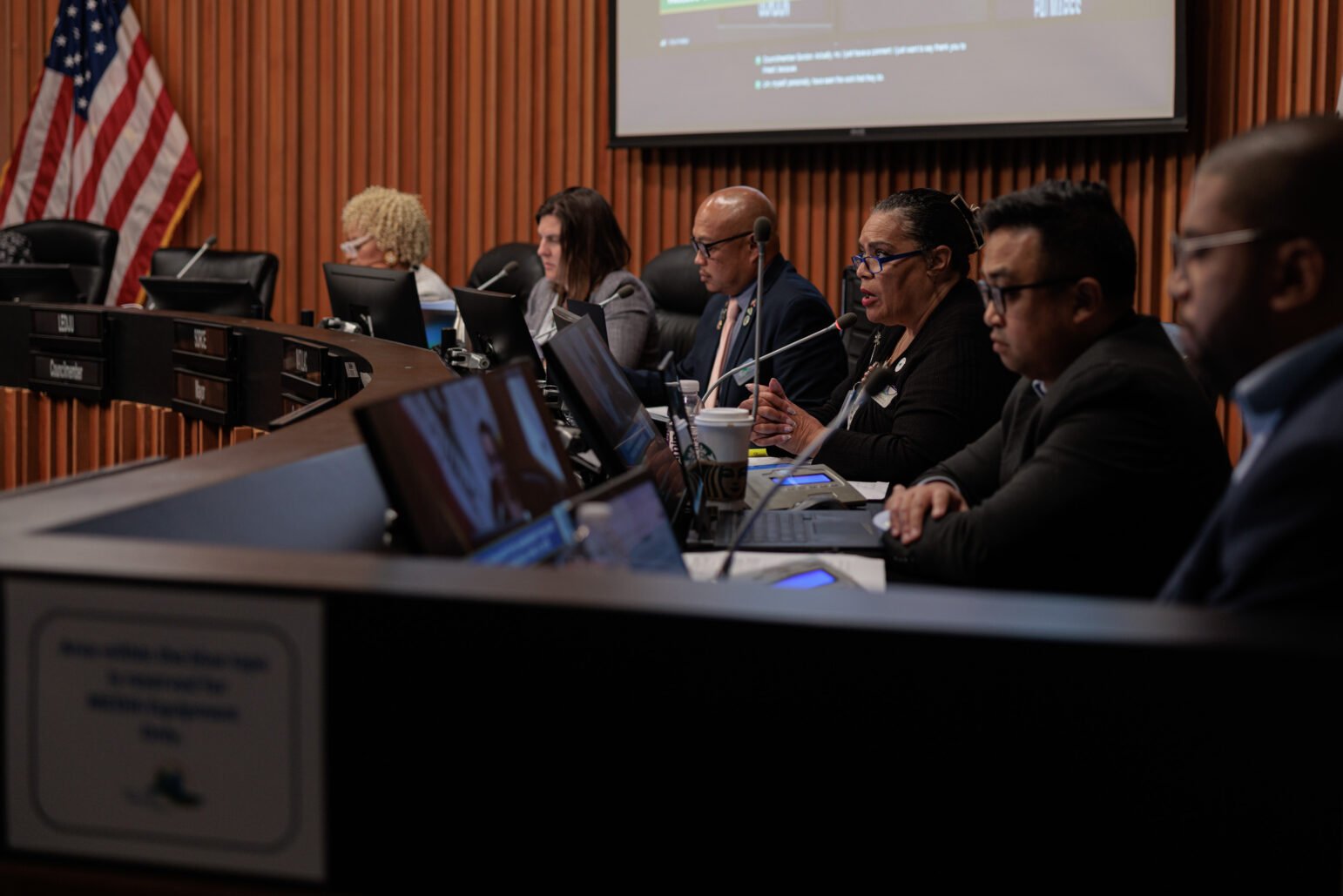 Vallejo City Council members sit at a curved dais with laptops and microphones during a public meeting, with an American flag and presentation screen in the background.