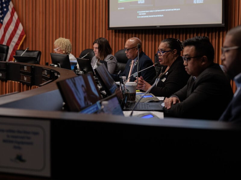 Vallejo City Council members sit at a curved dais with laptops and microphones during a public meeting, with an American flag and presentation screen in the background.