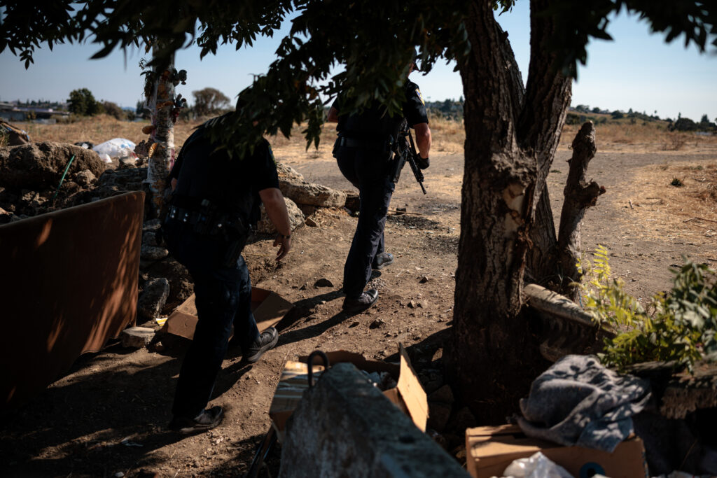 Two police officers move cautiously through a tree-lined area toward an open field, one carrying a rifle, during a daytime search.