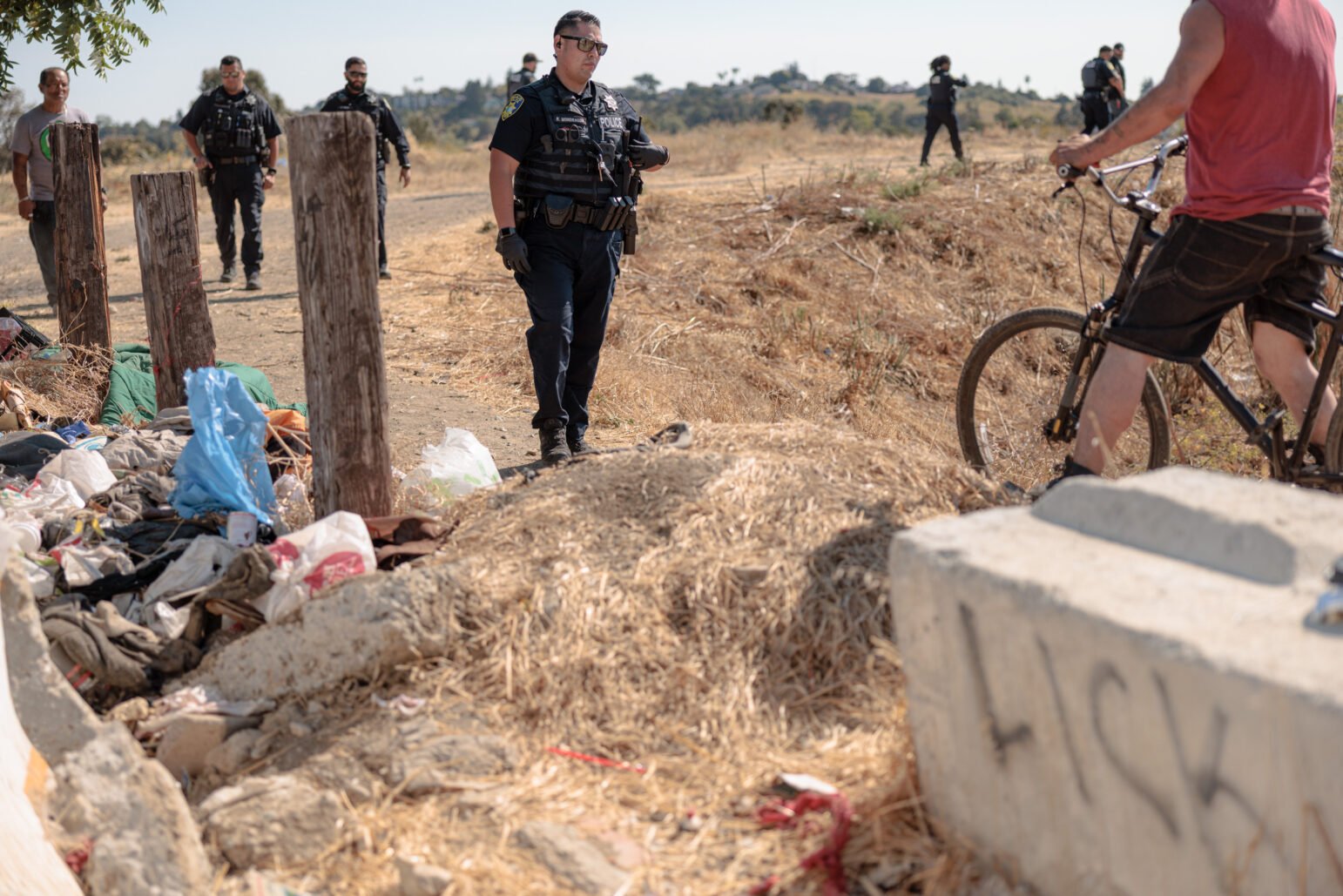 Several police officers walk through a dirt path lined with scattered belongings, as a man with a bicycle and another man stand nearby at an encampment.