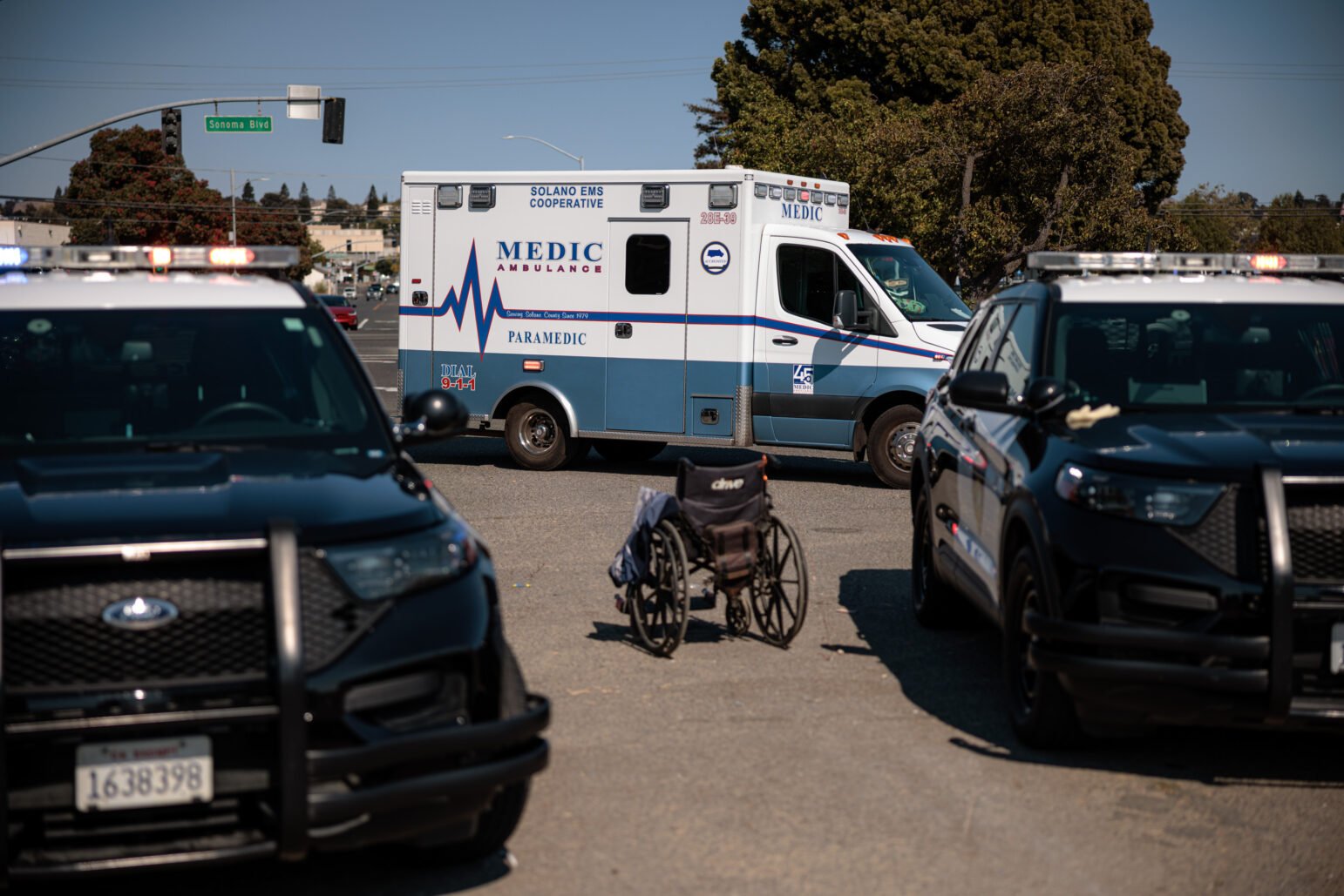 An ambulance is parked on a street near Sonoma Boulevard, flanked by two police SUVs, with an empty wheelchair positioned in the middle of the scene.
