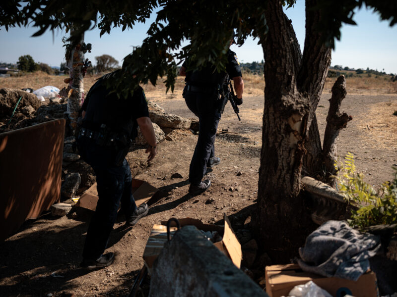 Two police officers move cautiously through a tree-lined area toward an open field, one carrying a rifle, during a daytime search.