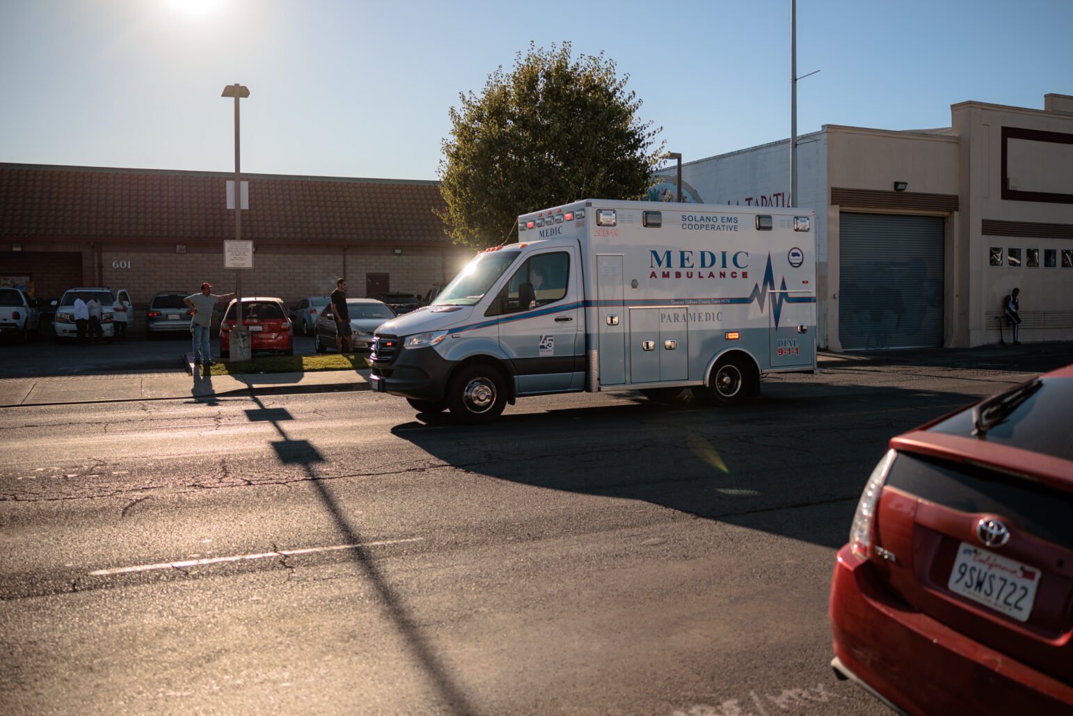 An ambulance drives down a street on a sunny street beside parked cars, with a few bystanders on the sidewalk.