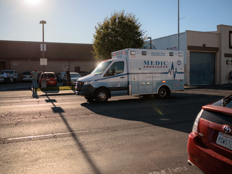 An ambulance drives away on a street, the sun glinting off its windshield, with a few bystanders on the sidewalk.