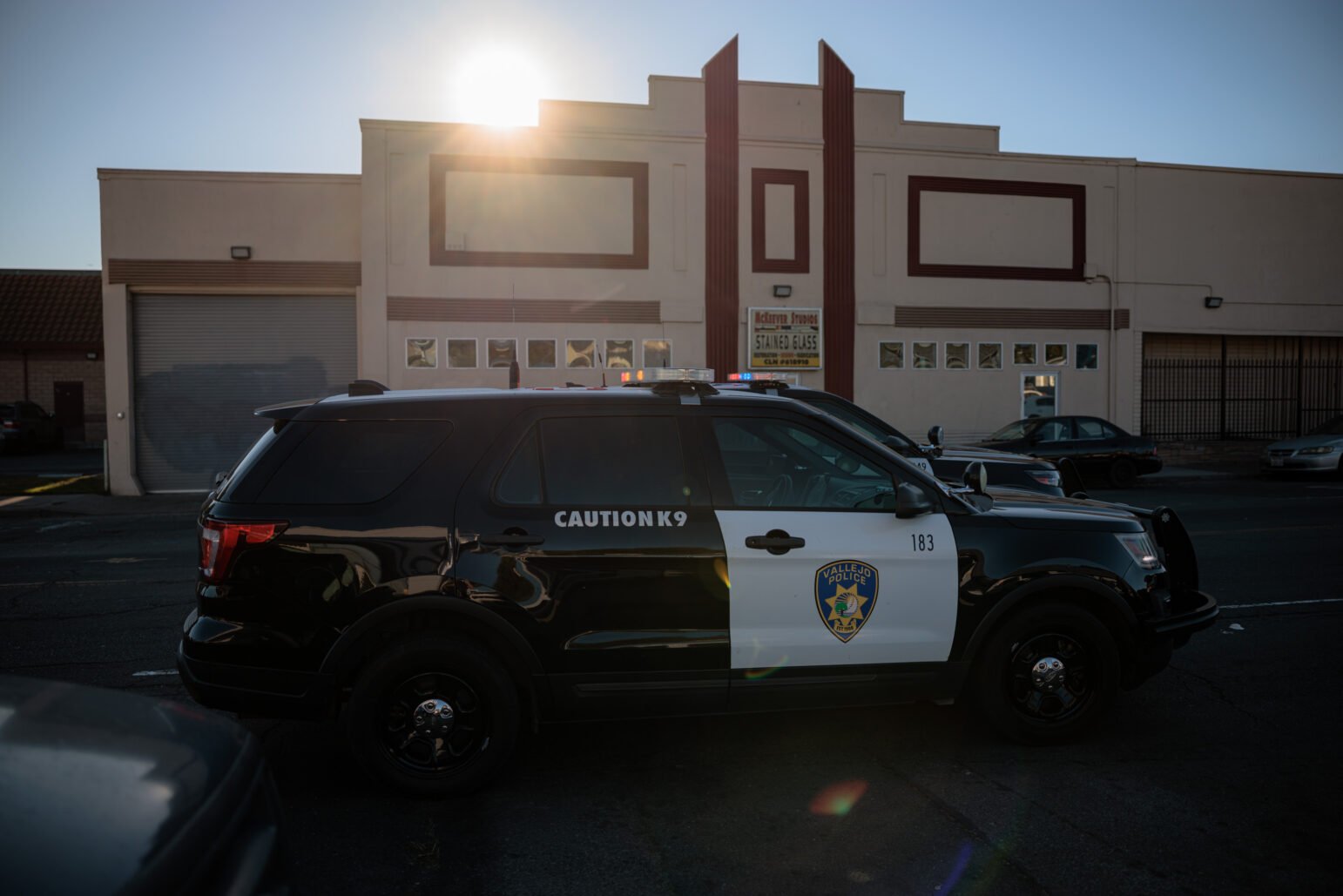 Two Vallejo Police Department SUVs parked in front of a beige storefront, backlit by the setting sun; “CAUTION K9” visible on the rear door of the nearest SUV.