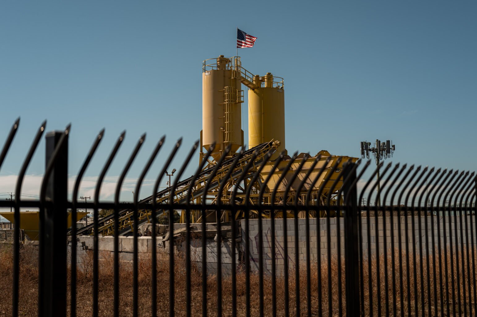 Through fence bars, an industrial site with two yellow silos and an angled conveyor; a U.S. flag flies above a cinderblock wall with, dry brush, clear sky.