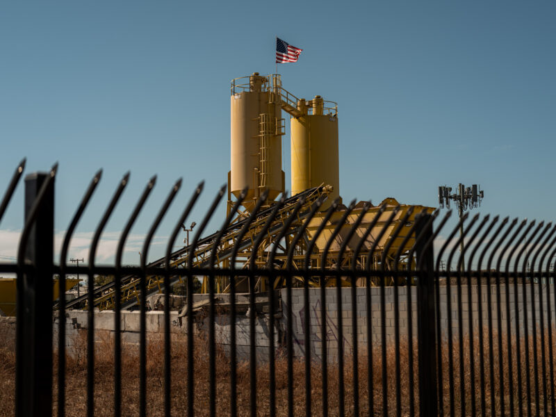 Through fence bars, an industrial site with two yellow silos and an angled conveyor; a U.S. flag flies above a cinderblock wall with, dry brush, clear sky.