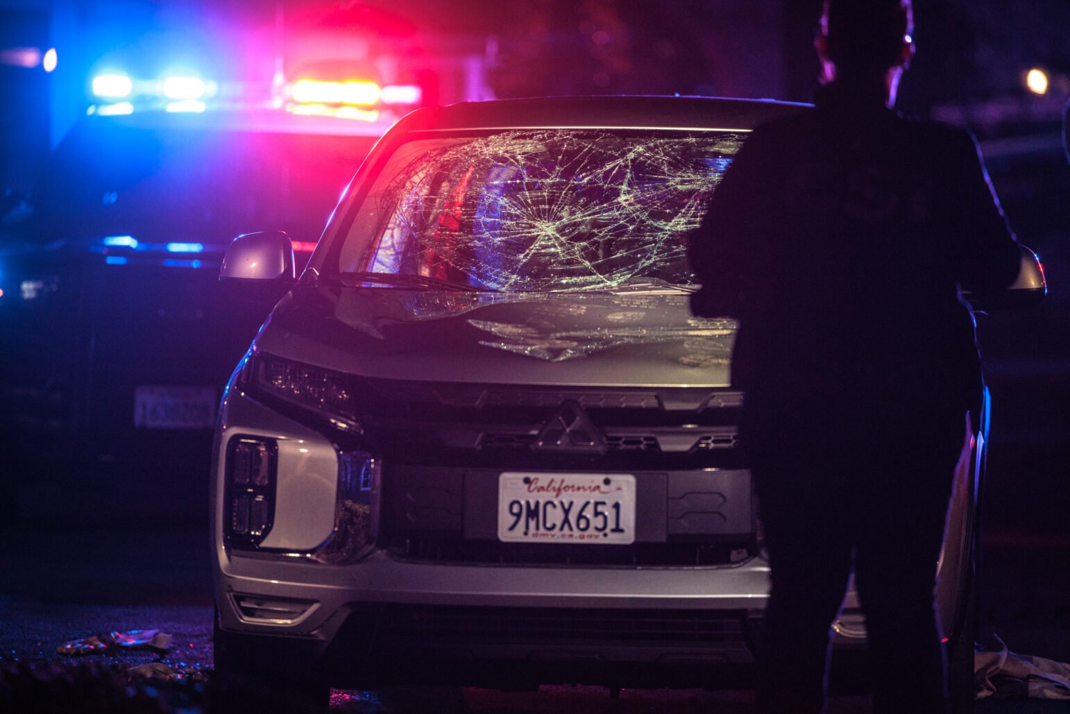 At night, a silver SUV with a spiderweb-shattered windshield sits in front of a police car with flashing red and blue lights; a person stands in silhouette at right.
