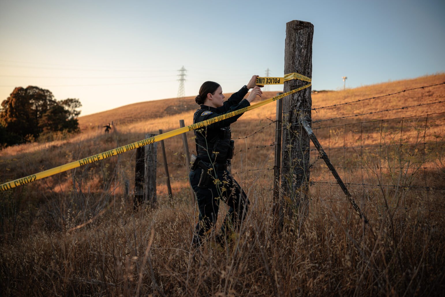Uniformed police officer secures yellow ‘POLICE LINE DO NOT CROSS’ tape to a wooden post along a barbed-wire fence in a dry hillside field at sunset, with distant power lines.