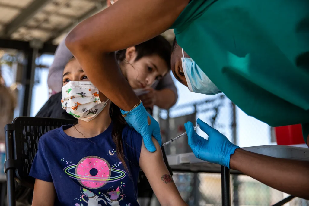 Child wearing a mask and a T-shirt with a donut design receives a vaccine injection in the arm from a healthcare worker in gloves and a mask.