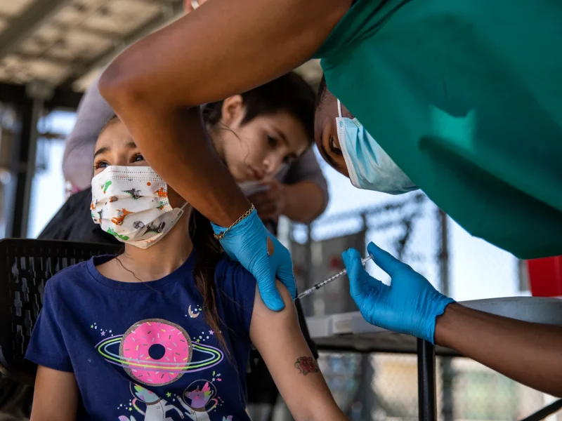 Child wearing a mask and a T-shirt with a donut design receives a vaccine injection in the arm from a healthcare worker in gloves and a mask.
