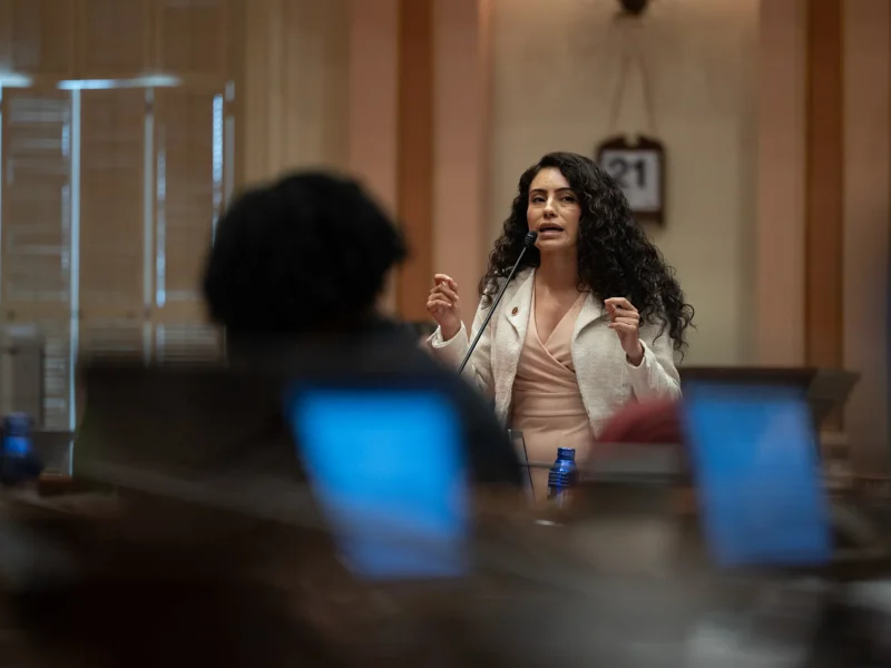 A woman with long curly hair in a light blazer speaks into a desk microphone in a legislative chamber, gesturing; blurred laptops and a blue water bottle frame the foreground.