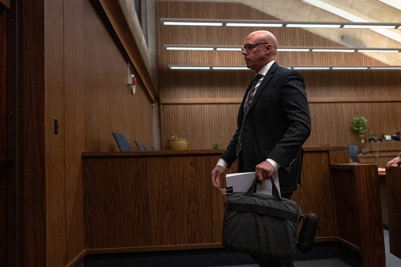 Bald man in a dark suit walks through a wood-paneled courtroom carrying a gray briefcase and paperwork, with fluorescent lights above and empty blue chairs in the background.