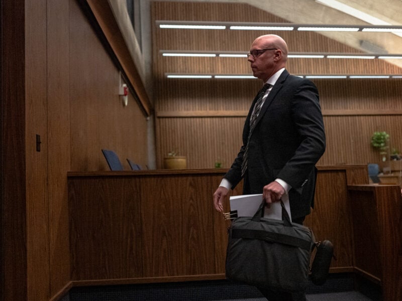 Bald man in a dark suit walks through a wood-paneled courtroom carrying a gray briefcase and paperwork, with fluorescent lights above and empty blue chairs in the background.